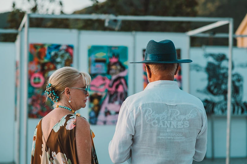 A sophisticated couple, the man in a stylish dark blue hat and the woman in an elegant dress, admire vibrant, avant-garde artworks at the Rubix Festival exhibition.