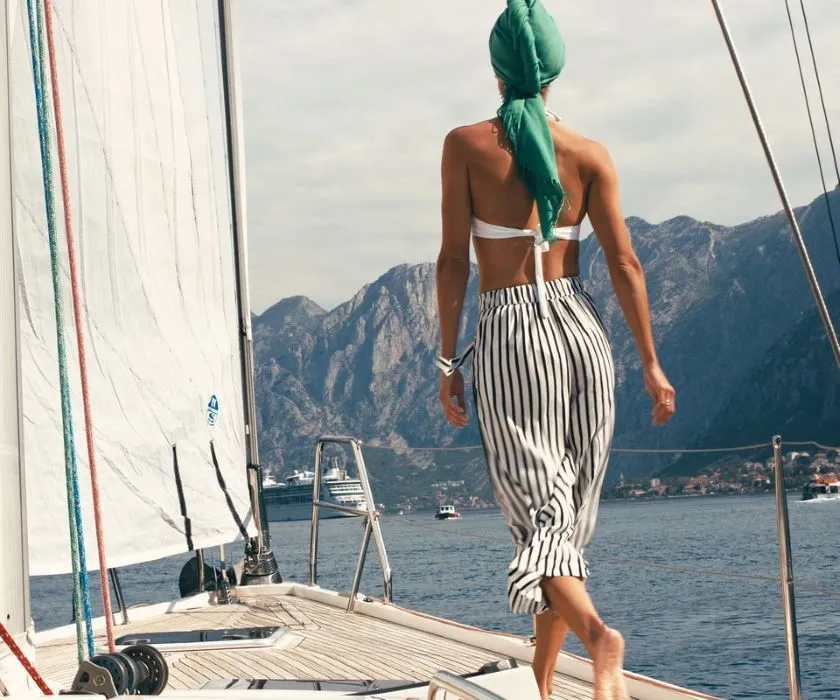 A woman in a striped outfit and green headscarf walking on a sailboat deck with mountains in the background.