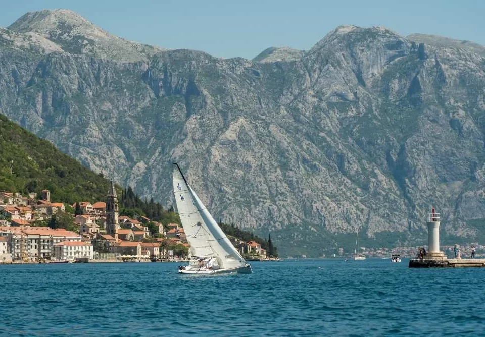 A sailboat gliding through the Bay of Kotor near the historic town of Perast, Montenegro, with dramatic mountain scenery and a lighthouse in the background.