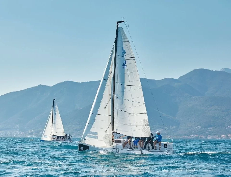 Two sailboats racing in the Porto Montenegro Yacht Club's Sailing Squadron, gliding across the deep blue waters with a stunning mountainous backdrop under clear skies.
