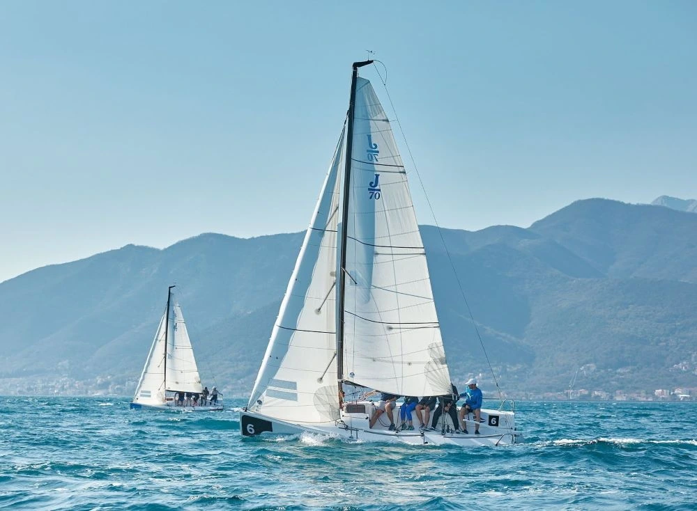 Two sailboats racing in the Porto Montenegro Yacht Club's Sailing Squadron, gliding across the deep blue waters with a stunning mountainous backdrop under clear skies.