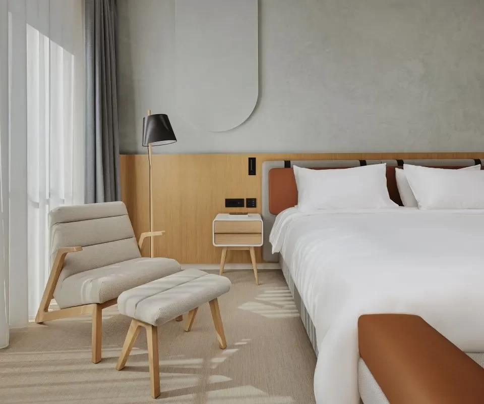 Cozy bedroom featuring a large bed with white bedding, a modern armchair with an ottoman, and a small wooden nightstand. The space is filled with natural light from full-length windows covered with soft curtains.
