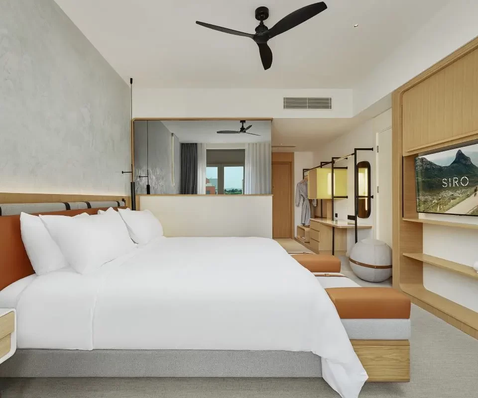 Spacious bedroom with a large bed in neutral tones, a ceiling fan, and a sleek TV mounted on a wooden entertainment unit. The design includes open wardrobe shelving, modern lighting, and a calm, minimalist aesthetic.