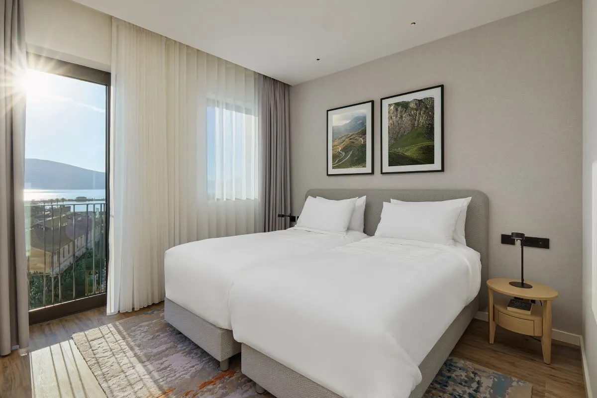 Bright twin bedroom with two neatly made beds, soft white bedding, and wall art depicting landscapes. Floor-to-ceiling windows and sheer curtains frame a breathtaking sea view with sunlight streaming in.