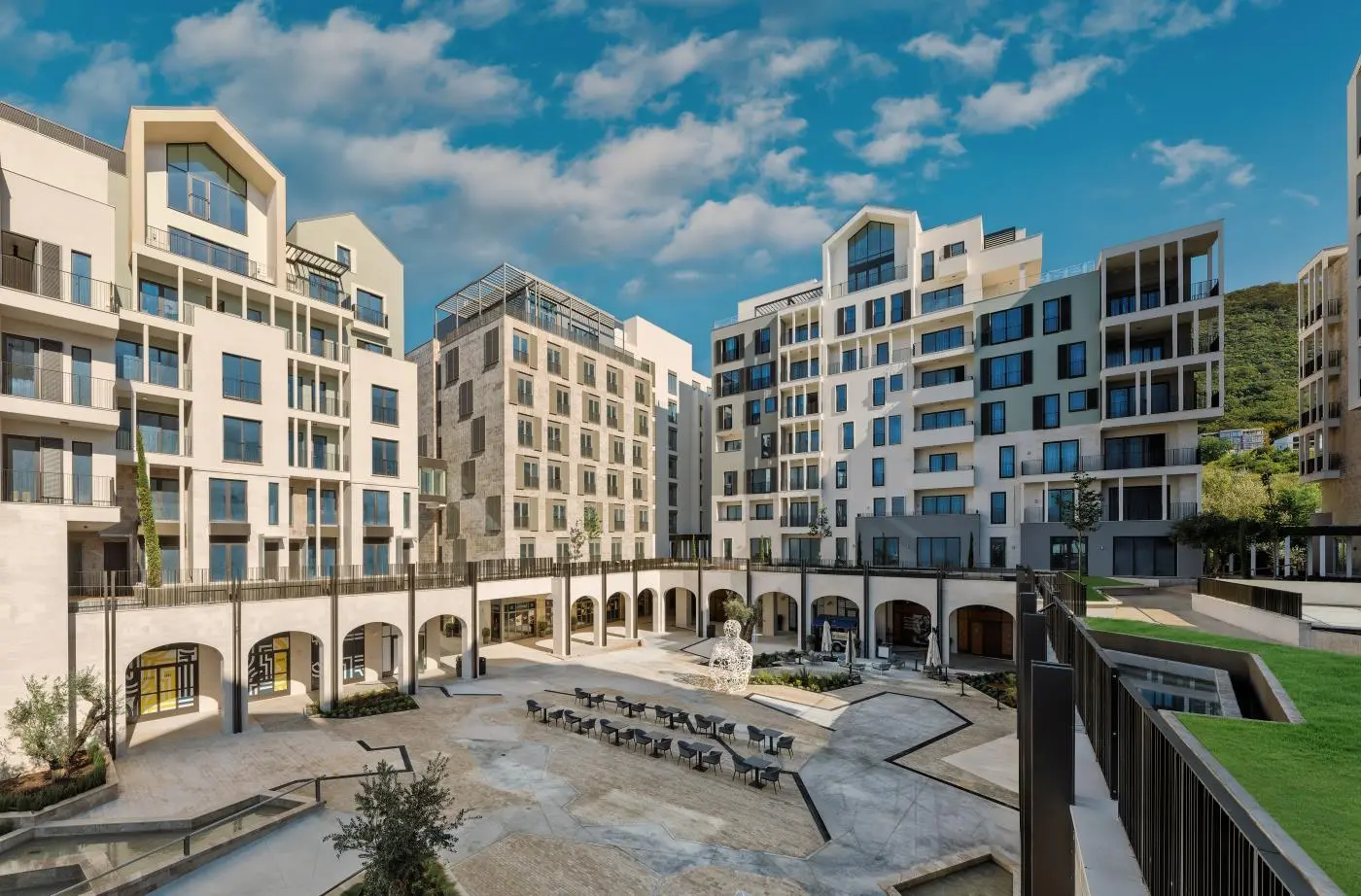 Modern Mediterranean-style residential and commercial buildings surrounding a spacious plaza with outdoor seating, architectural arches, and a centerpiece sculpture, set against a backdrop of greenery and a clear blue sky, Boka Place neighborhood.
