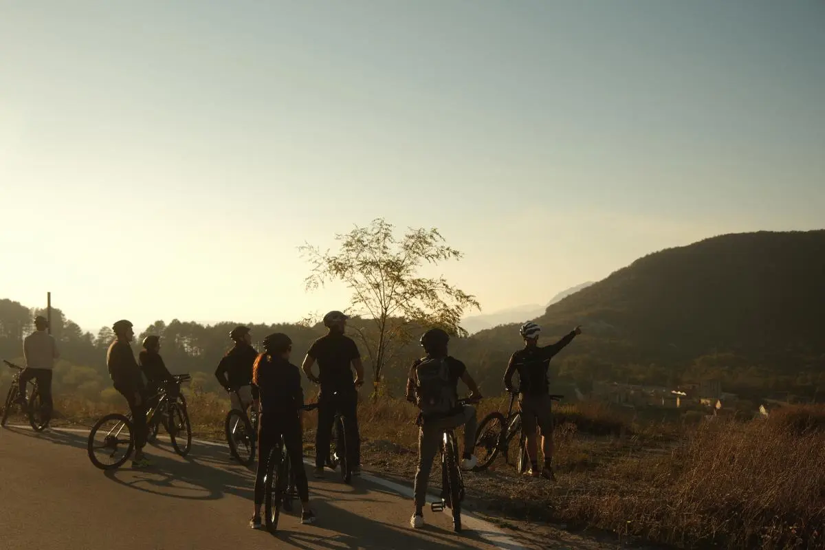 A group of cyclists pausing on a scenic roadside at sunset, with a guide pointing towards the horizon, surrounded by rolling hills and a peaceful rural landscape.