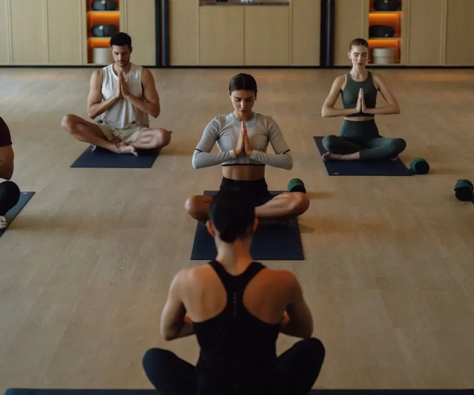 Group yoga class in a modern studio, with participants seated on mats practicing meditation. The space has wooden flooring and neatly organized fitness equipment in the background.