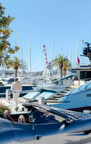 A person in light-colored clothing walks along a marina lined with luxury yachts, with a sleek convertible sports car in the foreground. Palm trees and sailboat masts rise against a clear blue sky, creating an atmosphere of affluence and leisure.