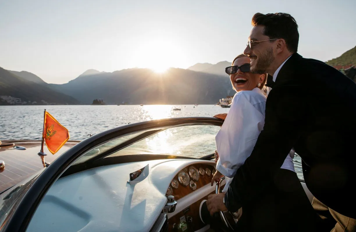 Stylish couple enjoying a luxury boat ride at sunset, with the man steering and the woman laughing, framed by the scenic backdrop of mountains and calm waters, symbolizing prosperity and the joy of exclusive experiences.