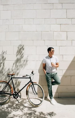 A fashionable man leans casually against a sunlit stone wall, holding a coffee cup, with a vintage-style bicycle parked beside him. Shadows of nearby trees create a relaxed atmosphere, capturing a moment of leisure and style in an urban setting.