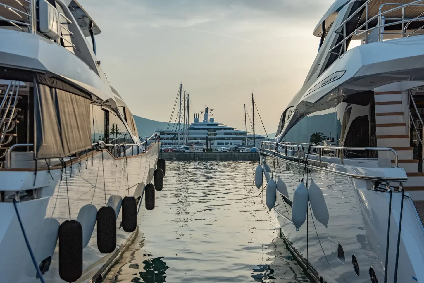 A stunning view between two luxurious yachts docked at Porto Montenegro Marina, with a grand superyacht in the distance, reflecting the opulence of the nautical lifestyle at sunset.