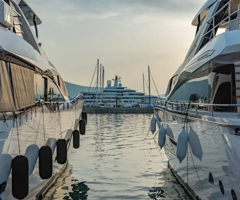A stunning view between two luxurious yachts docked at Porto Montenegro Marina, with a grand superyacht in the distance, reflecting the opulence of the nautical lifestyle at sunset.