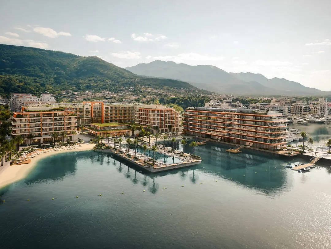 Luxurious waterfront residences stand majestically against a backdrop of lush, green mountains, with their contemporary architecture featuring expansive terraces. The Synchro Yards development is nestled beside a serene marina, offering direct access to the tranquil waters. A private beach with loungers and a large, palm-fringed pool deck completes the picture of an exclusive, upscale retreat.