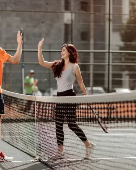 A young man in an orange sports club t-shirt and a young woman in a gray tank top high-five at the net after a tennis match on an outdoor court, with a cityscape and other players in the background.