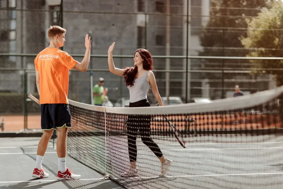 A young man in an orange sports club t-shirt and a young woman in a gray tank top high-five at the net after a tennis match on an outdoor court, with a cityscape and other players in the background.