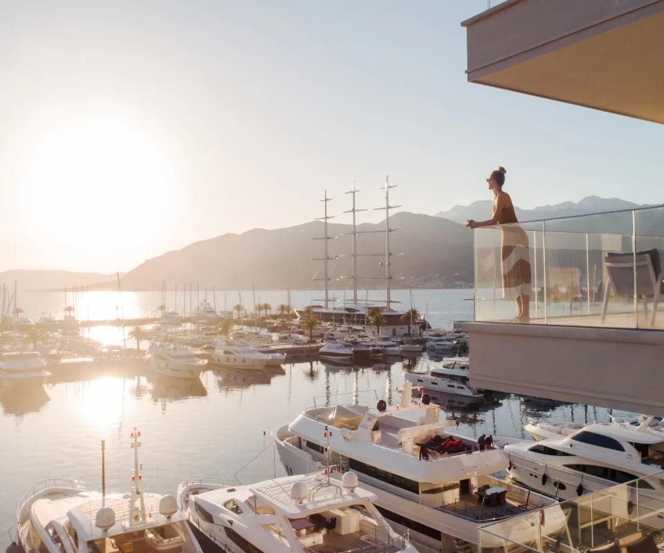 Woman standing on a luxurious balcony overlooking the marina in Porto Montenegro, basking in the serene sunset with yachts and sailboats docked below.