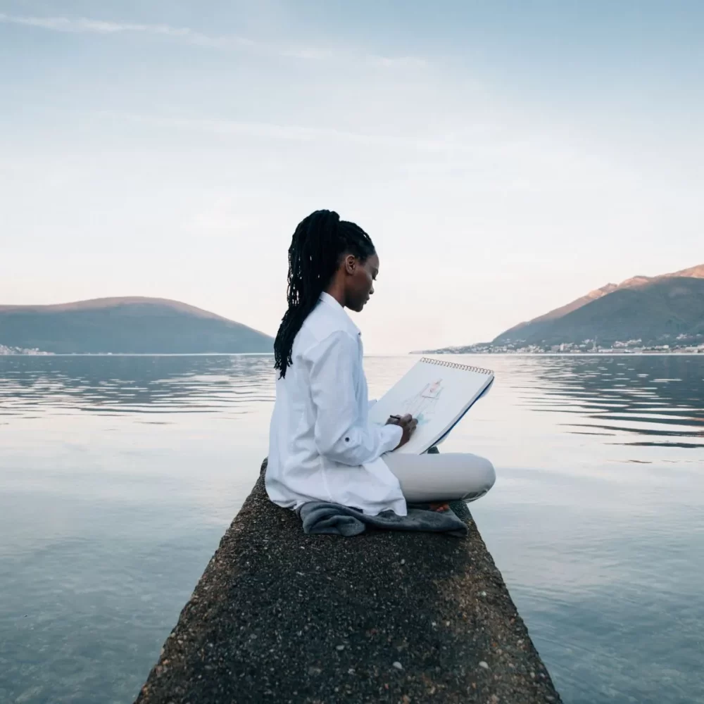 Woman sketching on a notepad while sitting on a pier overlooking a calm lake with mountains in the background.