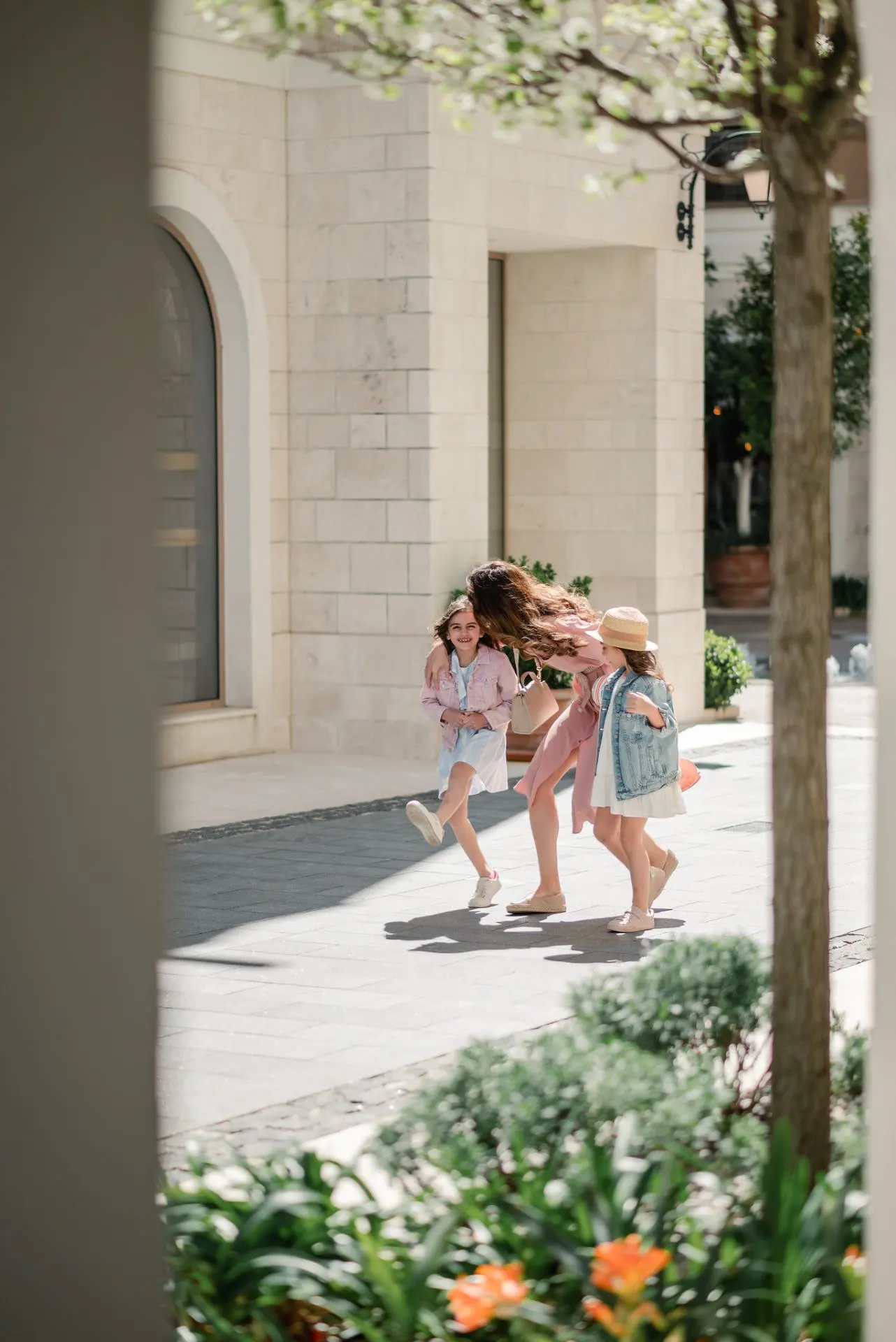 A stylishly dressed woman in a pink dress playfully walks with two young girls, both in light summer outfits, along a sunlit, upscale stone-paved street lined with lush greenery and elegant buildings, evoking a sense of joy and luxury.