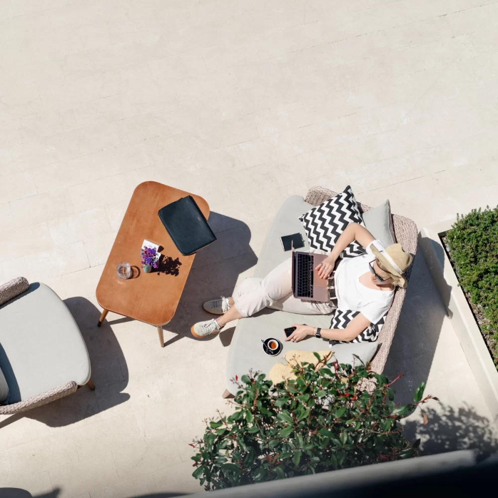 Woman working on a laptop while relaxing on an outdoor patio with a coffee and a tablet on a table.