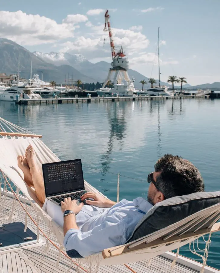 A man wearing sunglasses and a light blue shirt is lying in a hammock on the deck of a yacht, working on his laptop. The marina is surrounded by calm waters, snow-capped mountains, palm trees, and a large industrial crane in the background.