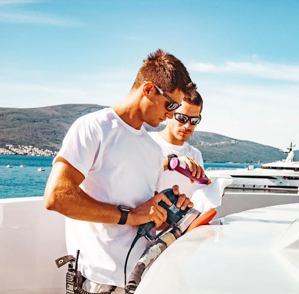 Two young men wearing sunglasses and white t-shirts are polishing a yacht's surface under bright sunlight. One is using an electric polisher, while the other applies a cleaning product with a cloth. The background features calm blue waters, a coastal town, and luxury yachts docked nearby.