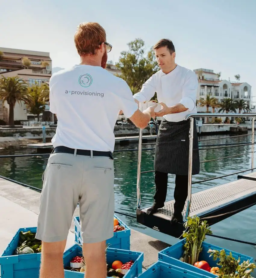 A provisioning service worker wearing a white t-shirt with the "a+ provisioning" logo hands a package to a yacht chef dressed in a white uniform and a striped apron. They are standing on a marina dock with blue crates filled with fresh vegetables and fruits. In the background, there are luxury waterfront buildings with palm trees.