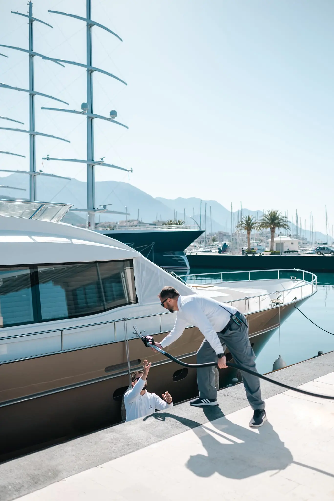 A yacht crew members in Porto Montenegro Marina passing a fuel hose to a colleague on board, with luxury yachts and towering sailboats moored in the background, set against a scenic mountain view.