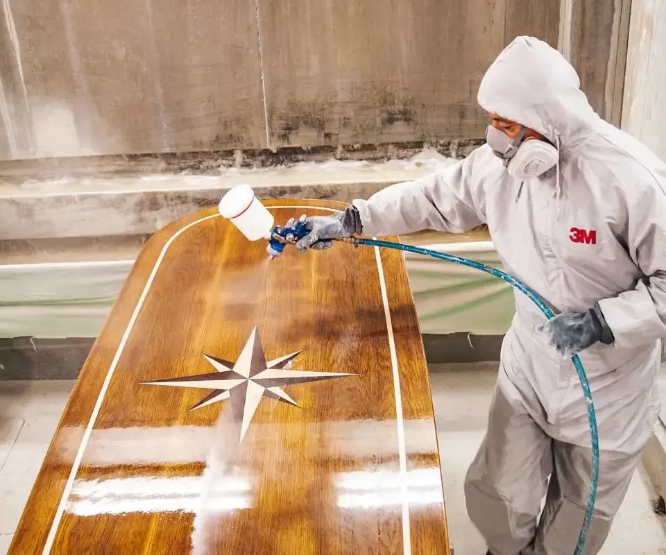 A worker in a protective 3M suit, gloves, and a respirator mask is spray-coating a high-gloss varnish onto a wooden yacht table with a compass rose inlay. The setting appears to be a professional workshop or shipyard, with an industrial environment in the background.
