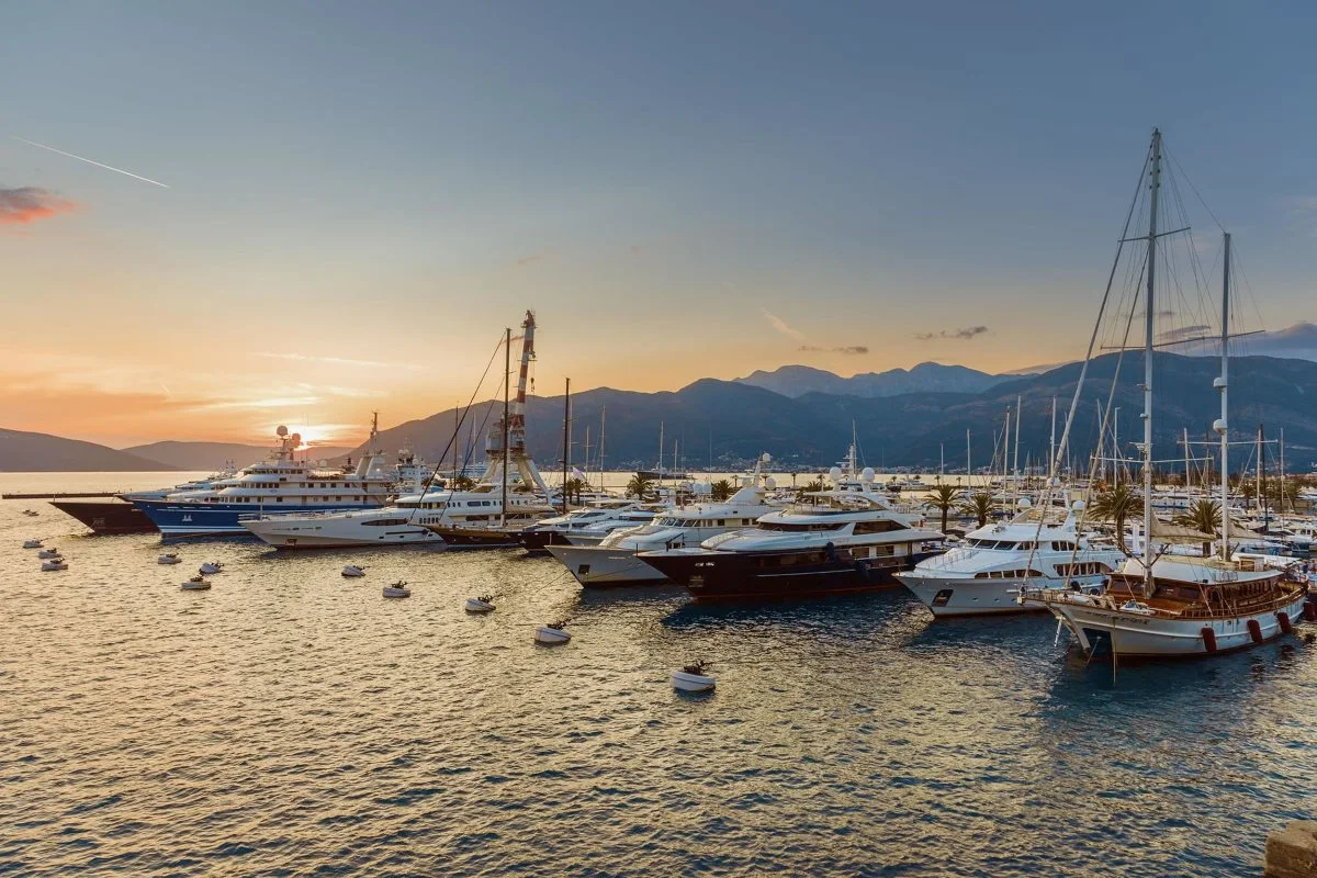 A serene sunset over Porto Montenegro Marina, showcasing luxurious yachts moored against a backdrop of mountains and calm waters, reflecting the golden hues of the sky.