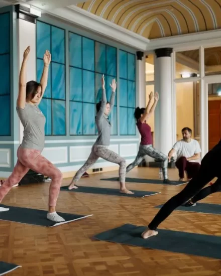 A group of women in athletic wear participate in a yoga session inside a beautifully decorated ballroom with high ceilings, blue paneling, and wooden flooring, stretching their arms upwards in a warrior pose.