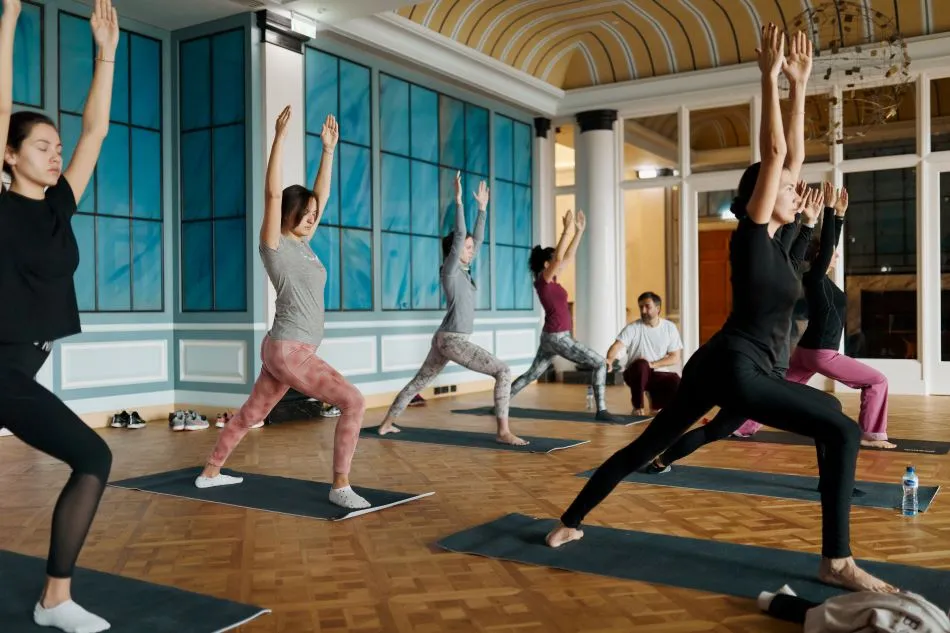 A group of women in athletic wear participate in a yoga session inside a beautifully decorated ballroom with high ceilings, blue paneling, and wooden flooring, stretching their arms upwards in a warrior pose.