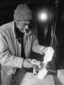 Black and white portrait of Zoran Obrenovic working with a power tool, wearing a knit hat and smoking a cigarette.